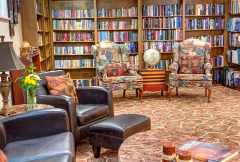 A cozy library or reading room with two patterned armchairs and two black leather chairs arranged around a small wooden table with a globe on it. The background features tall wooden bookshelves filled with books. The floor is carpeted with a patterned design, and there is a small table with a lamp and a vase of yellow flowers.