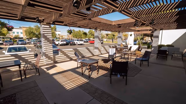 Outdoor covered patio area with wooden pergola casting striped shadows on the ground. Several chairs and tables are arranged under the pergola. In the background, there is a parking lot with multiple cars and some trees.