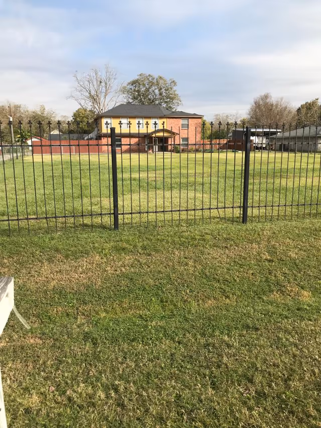 A large grassy yard enclosed by a black metal fence with a two-story brick and yellow building in the background under a partly cloudy sky.
