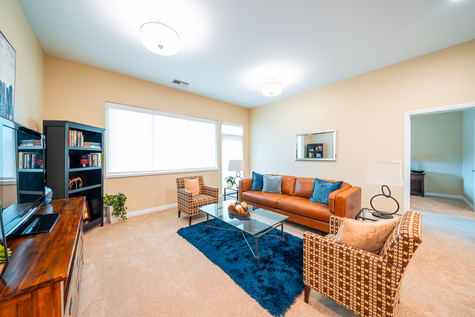 A bright living room with beige walls and carpet, featuring a brown leather sofa with blue and gray pillows, two patterned armchairs with beige cushions, a glass coffee table on a dark blue rug, a wooden TV stand with a flat-screen TV, a black bookshelf with books and decorative items, a floor lamp, and a large window with white blinds.