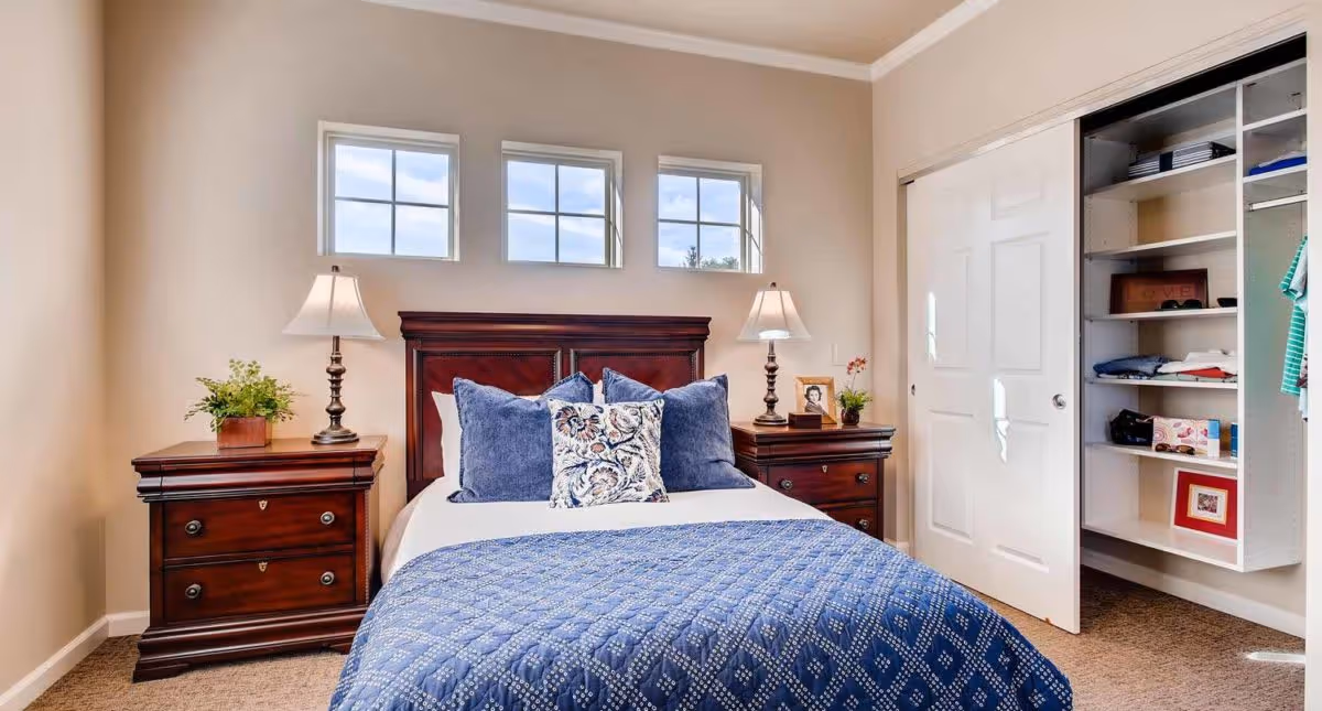 A neatly made bed with a dark wooden headboard and blue patterned bedding is centered between two matching wooden nightstands, each with a lamp and decorative items. Above the bed are three small square windows letting in natural light. To the right, there is an open closet with sliding white doors revealing shelves and hanging clothes.