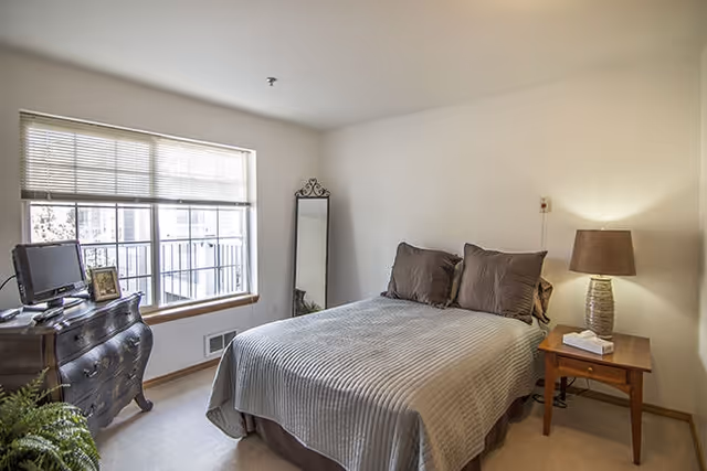 Sunlit bedroom with a double bed, bedside table and lamp, dresser with a small TV, floor mirror, and a large window.