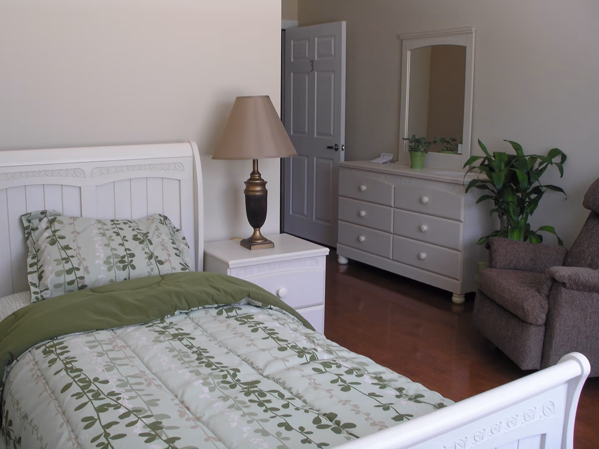 A senior care facility bedroom with a white wooden bed featuring green and beige leaf-patterned bedding. Next to the bed is a white nightstand with a bronze and black lamp. In the background, there is a white dresser with a mirror and a green potted plant on top. A brown upholstered recliner chair is positioned near the dresser, and a door marked with the number 3 is partially open.