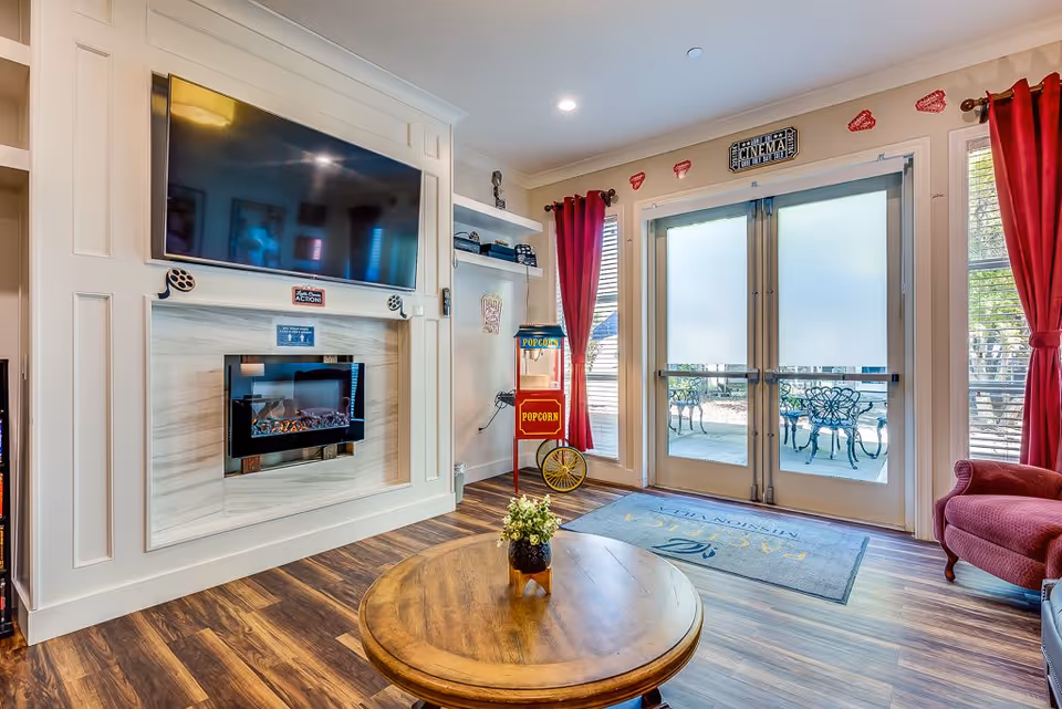 A cozy senior living common area featuring a wall-mounted flat screen TV above a modern electric fireplace. The room has wood flooring, a round wooden coffee table with a small plant, red curtains framing glass double doors leading to an outdoor patio with metal chairs and tables. A vintage-style popcorn machine stands near the doors, and decorative movie-themed signs adorn the walls.