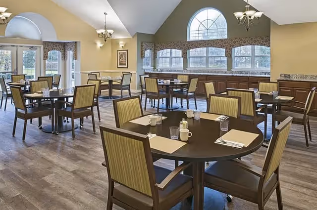 Well-lit dining room with multiple round tables and wooden chairs set with placemats and cups beneath arched windows and chandeliers.