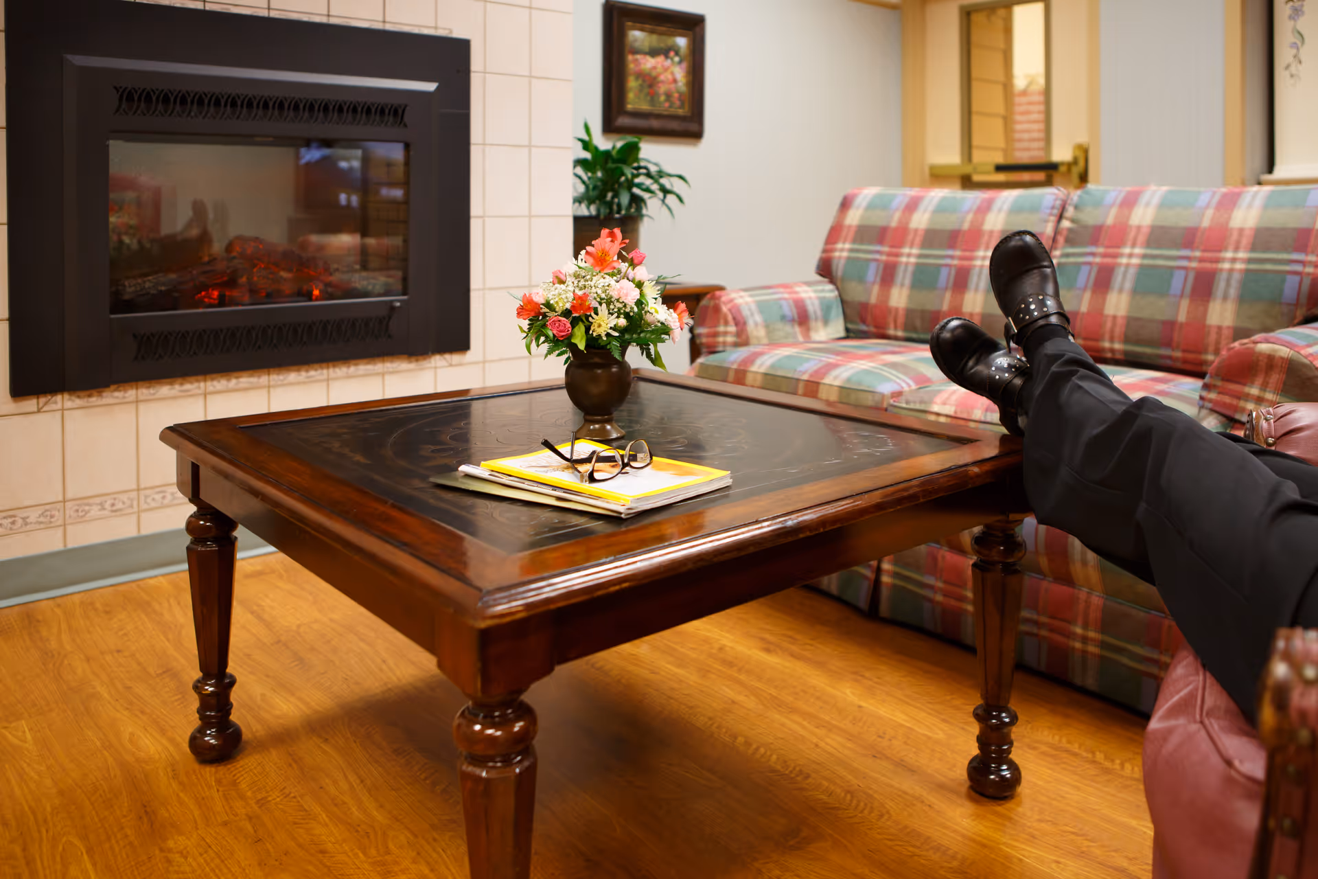 A cozy living room area with a wooden coffee table holding a vase of flowers and some reading materials. Behind the table is a plaid-patterned couch, and to the left is a wall-mounted electric fireplace. A person is sitting on a chair with their legs crossed, wearing black pants and black boots.