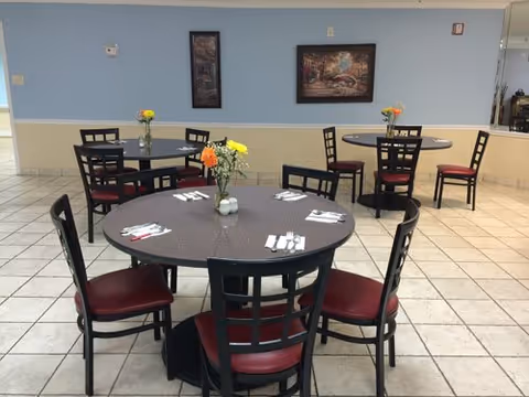 Dining area with round tables, chairs, place settings, and small floral centerpieces against a light blue wall.