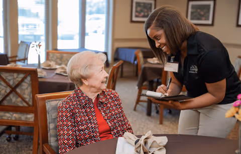 An elderly woman sitting at a dining table inside a facility, smiling and looking up at a staff member who is standing beside her, writing on a notepad. The room has multiple tables and chairs, with large windows in the background letting in natural light.
