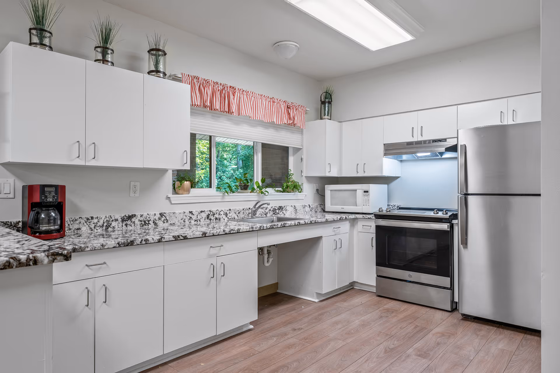 A clean and modern kitchen with white cabinets, a marbled countertop, a stainless steel refrigerator and stove, a white microwave, a red coffee maker, and several potted plants on the windowsill and above the cabinets. The kitchen has a window with a red and white striped valance and wood flooring.