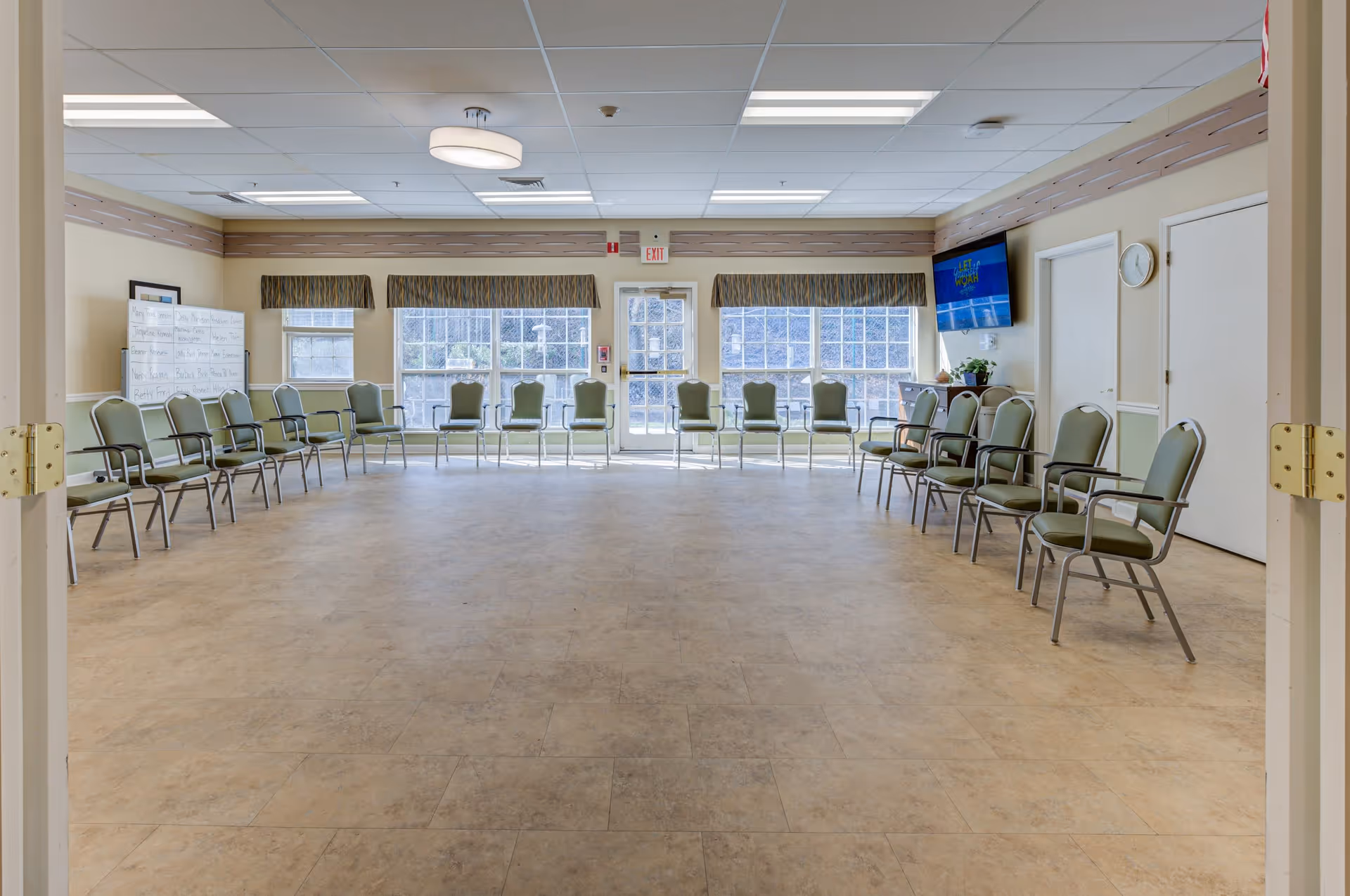 A large, bright room with beige tiled flooring and light-colored walls. The room is arranged with green cushioned chairs placed in a wide circle along the walls. There are three large windows and a glass door letting in natural light. A whiteboard with writing is visible on the left side, and a wall-mounted TV is on the right side above a small cabinet. A clock is mounted on the wall near two closed doors.