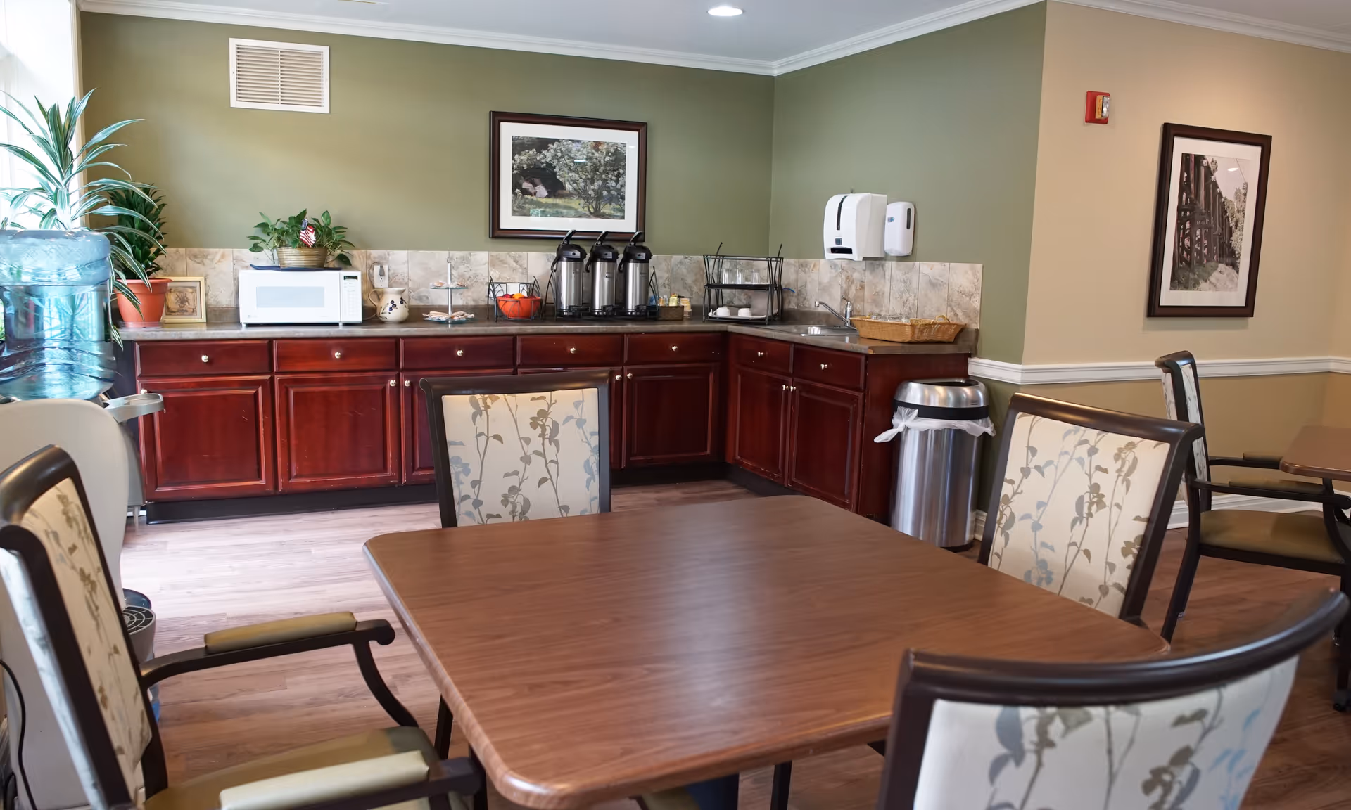 A communal dining area with a wooden table and upholstered chairs in front of a kitchenette featuring dark wood cabinets, a microwave, coffee dispensers, and a water cooler.