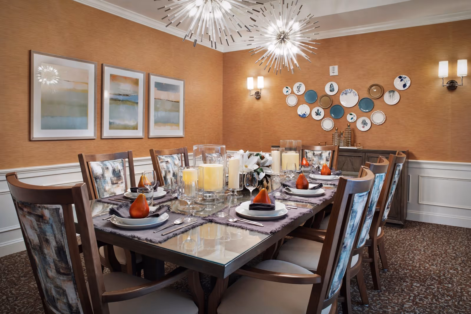 Elegant dining room with a long set table arranged with plates, candles and decorative pears, surrounded by upholstered chairs under modern chandeliers.