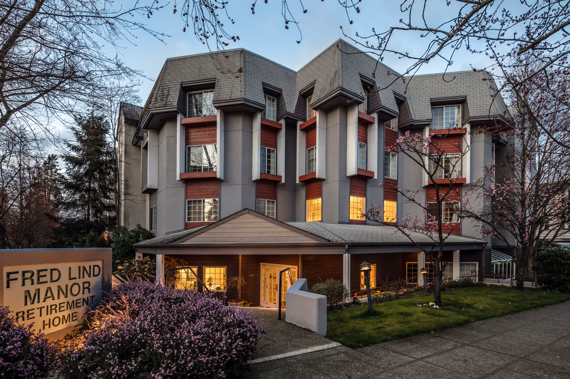 Exterior view of Fred Lind Manor retirement home building at dusk, showing a multi-story structure with lit windows, surrounded by trees and bushes with blooming flowers, and a sign in front reading 'Fred Lind Manor Retirement Home'.