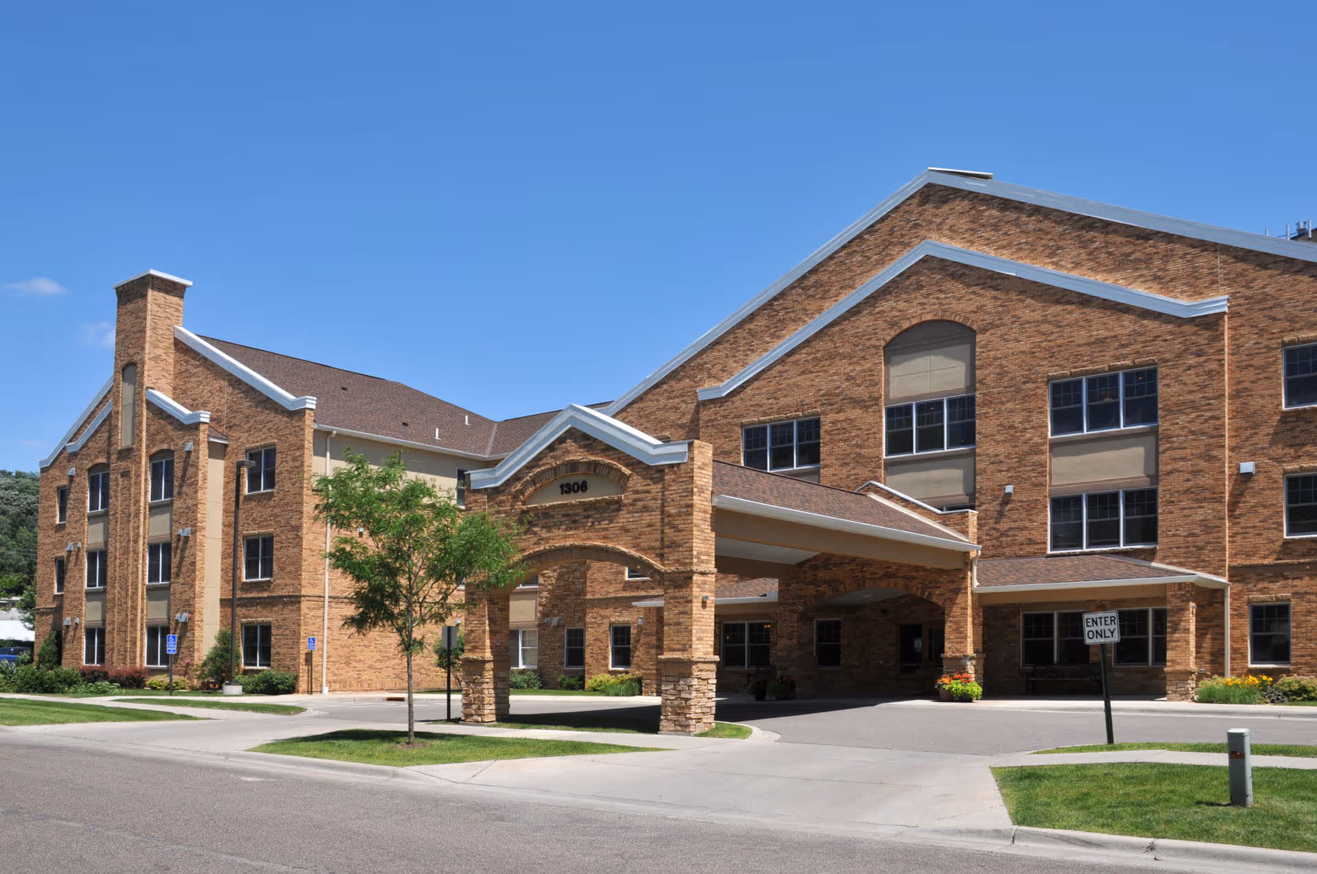 Front exterior of a three-story brick building with a covered porte-cochere and driveway beneath a clear blue sky.