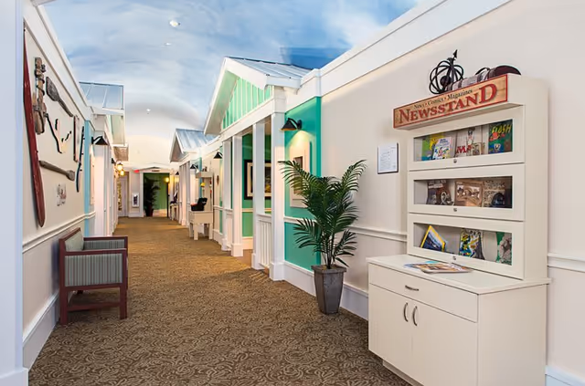 A brightly lit indoor hallway designed to resemble an outdoor street scene with small house-like facades painted in white and green. The hallway features a carpeted floor, a bench on the left side, a potted plant, and a white newsstand cabinet displaying magazines and comics on the right side. The ceiling is painted to look like a blue sky with clouds.