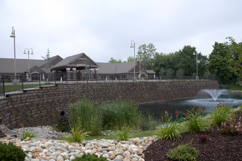 Outdoor view of a facility with a stone retaining wall, a pond with a water fountain, landscaped plants, and a building with a gray roof and multiple lamp posts in the background under a cloudy sky.