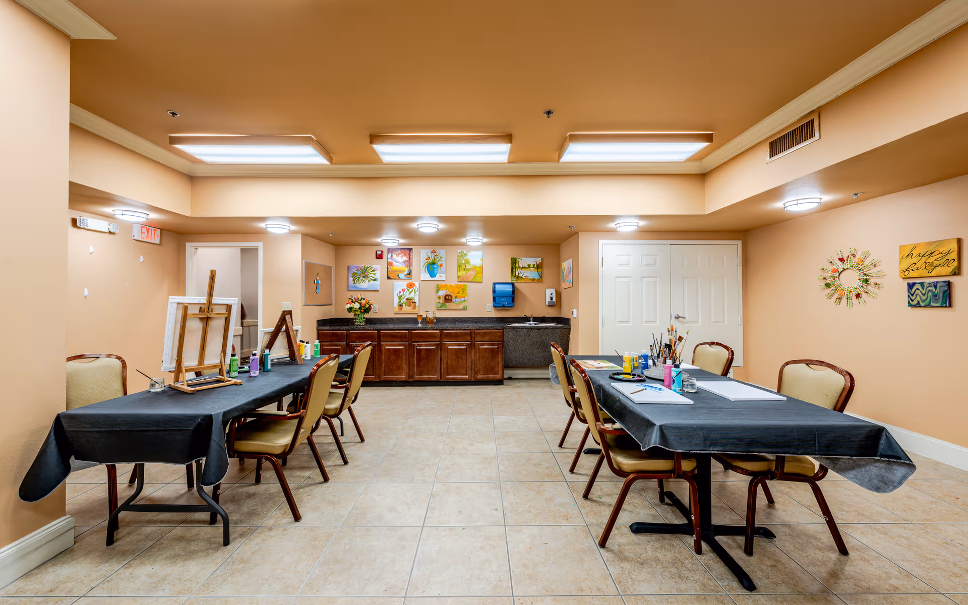 A well-lit activity room with two tables covered in black tablecloths, each surrounded by chairs. The tables have art supplies including paint bottles, brushes, and canvases on easels. The walls are decorated with colorful paintings and cheerful wall art. There is a countertop with cabinets and a sink at the back of the room.