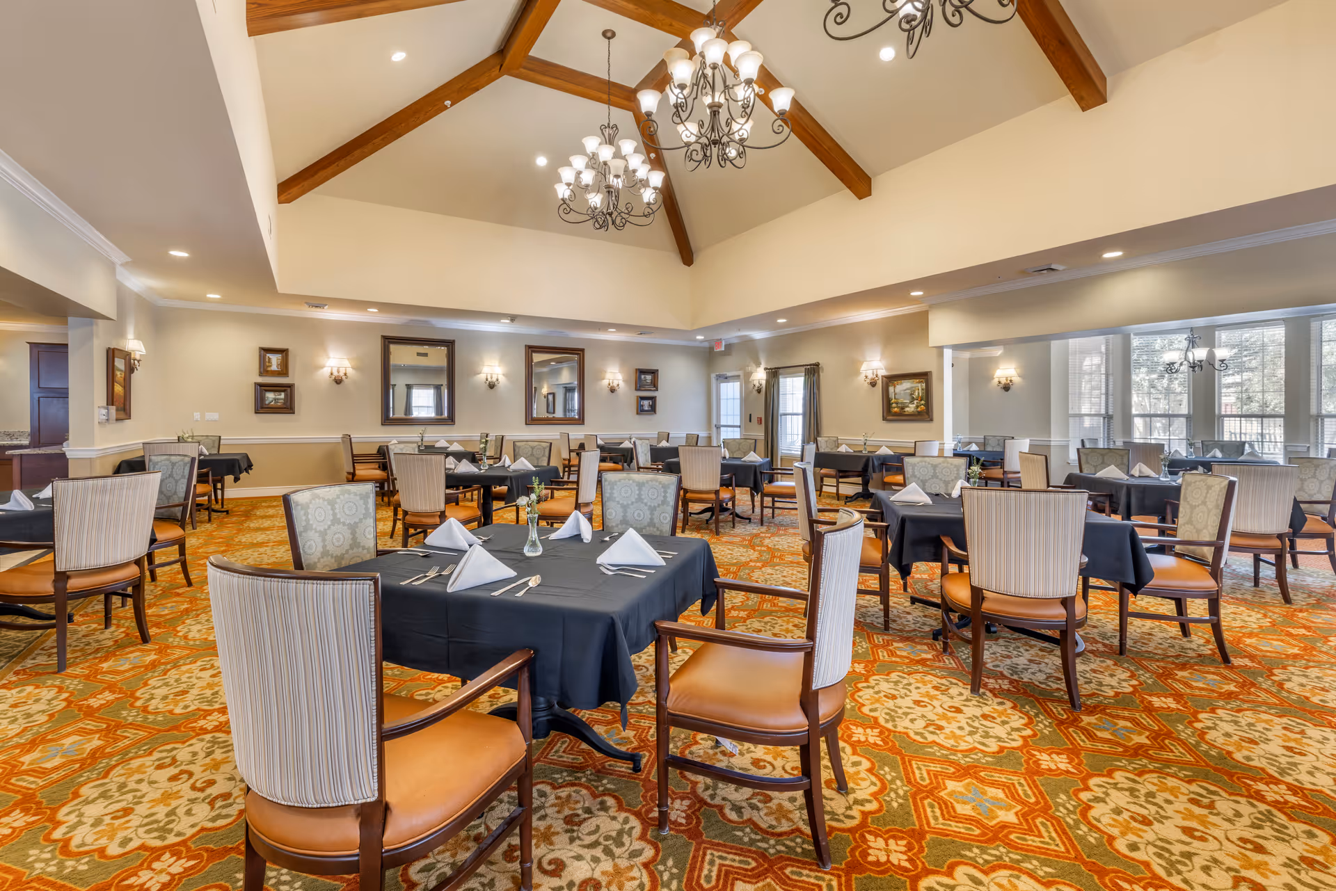 Spacious dining room with multiple tables set with black tablecloths and folded napkins beneath chandeliers and a vaulted ceiling.