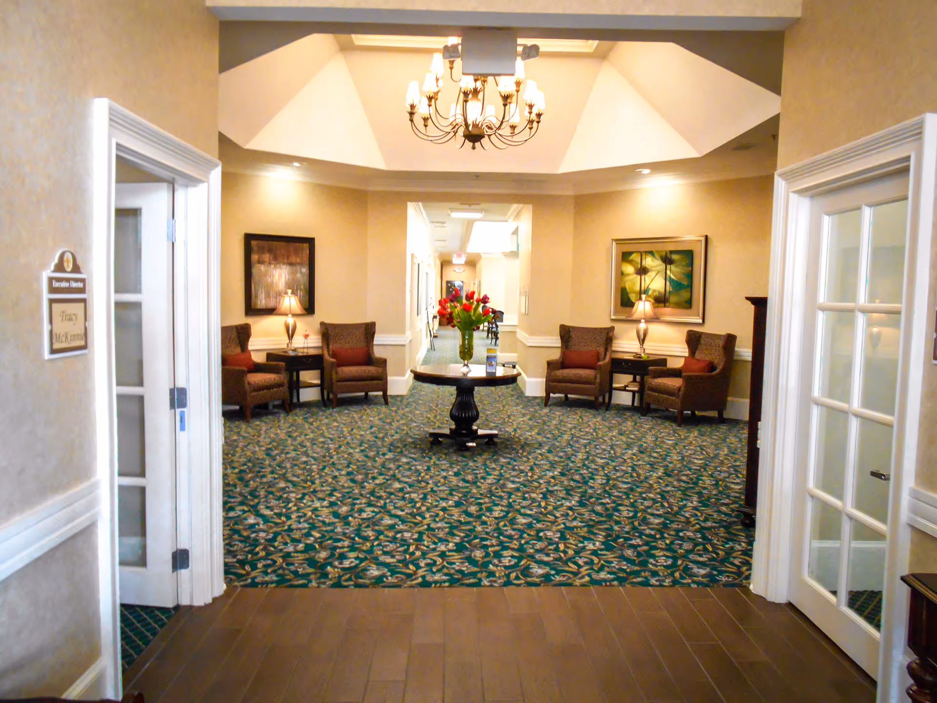 Entrance view into a decorated common lounge with a central table of flowers, chandelier, and upholstered seating.