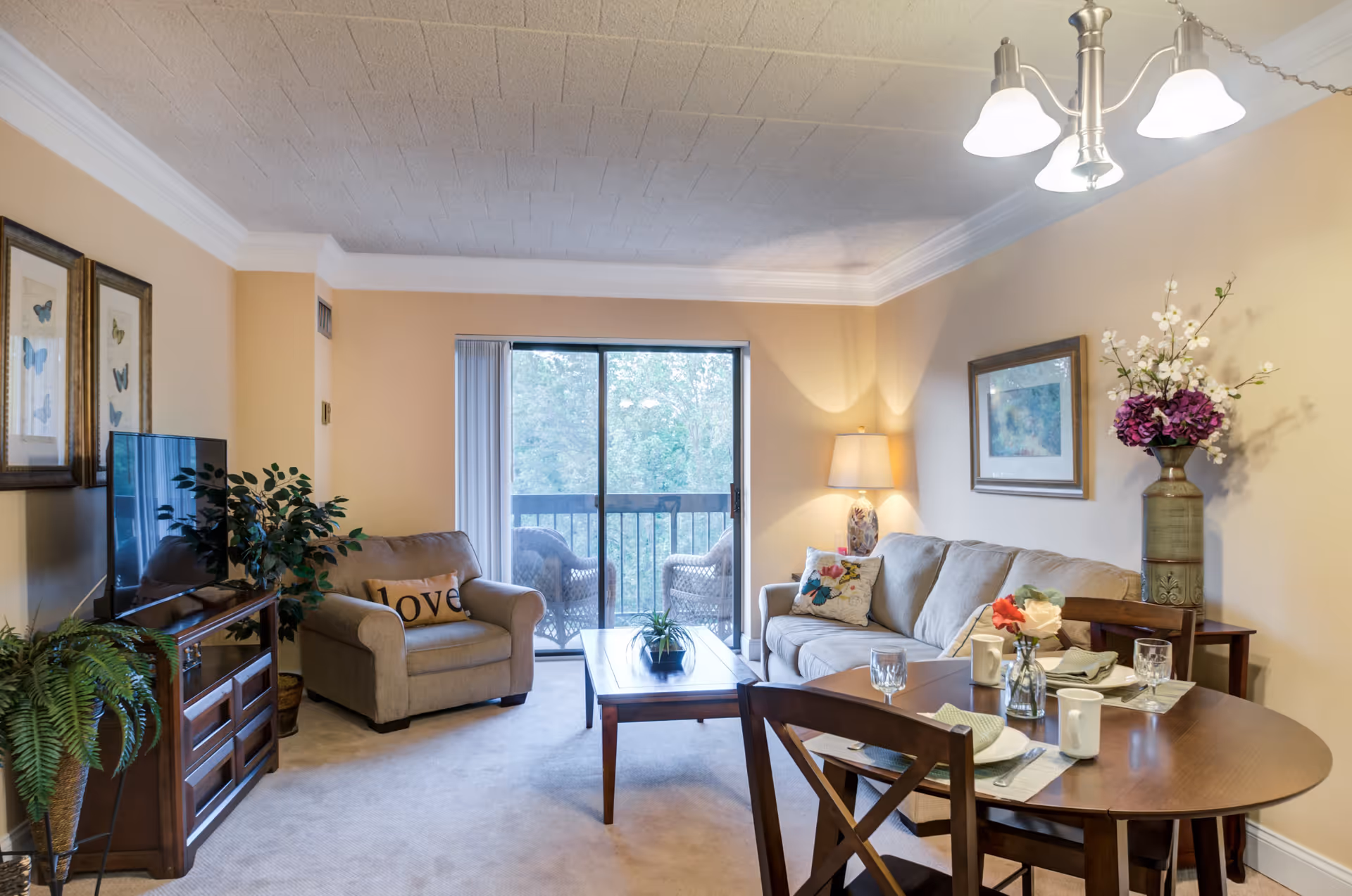 A cozy living room with beige walls and carpet, featuring a beige sofa with decorative pillows, a matching armchair with a 'love' pillow, a wooden coffee table, and a wooden dining table set with plates, glasses, and mugs. There is a TV on a wooden stand, framed butterfly artwork on the wall, a floor lamp, a large vase with flowers, and a sliding glass door leading to a balcony with two chairs and greenery outside.