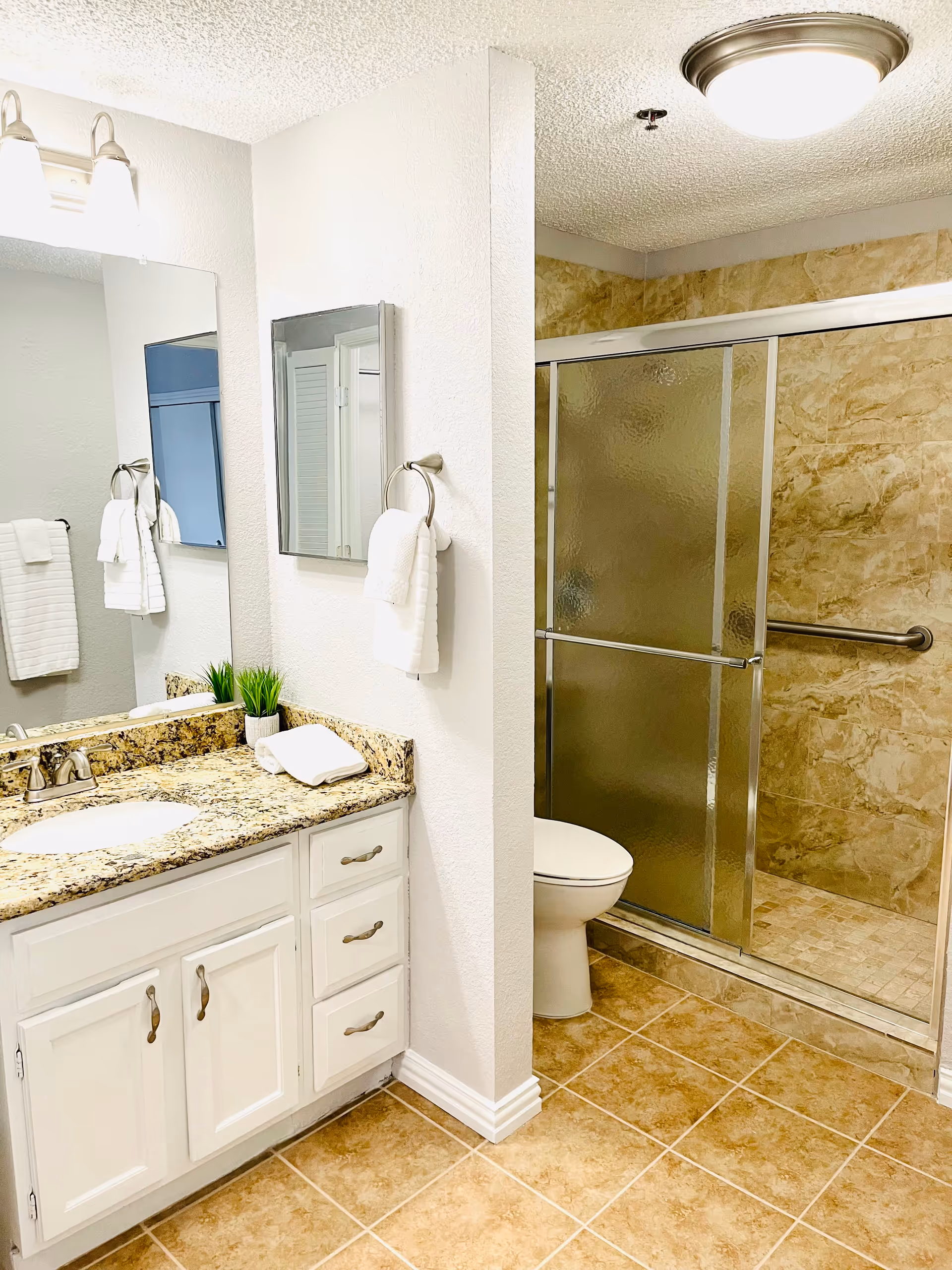 A clean and bright bathroom featuring a granite countertop with a sink, white cabinets, a mirror, and a towel ring with a white towel. The bathroom also includes a toilet and a shower with sliding glass doors and beige tiled walls and floor.