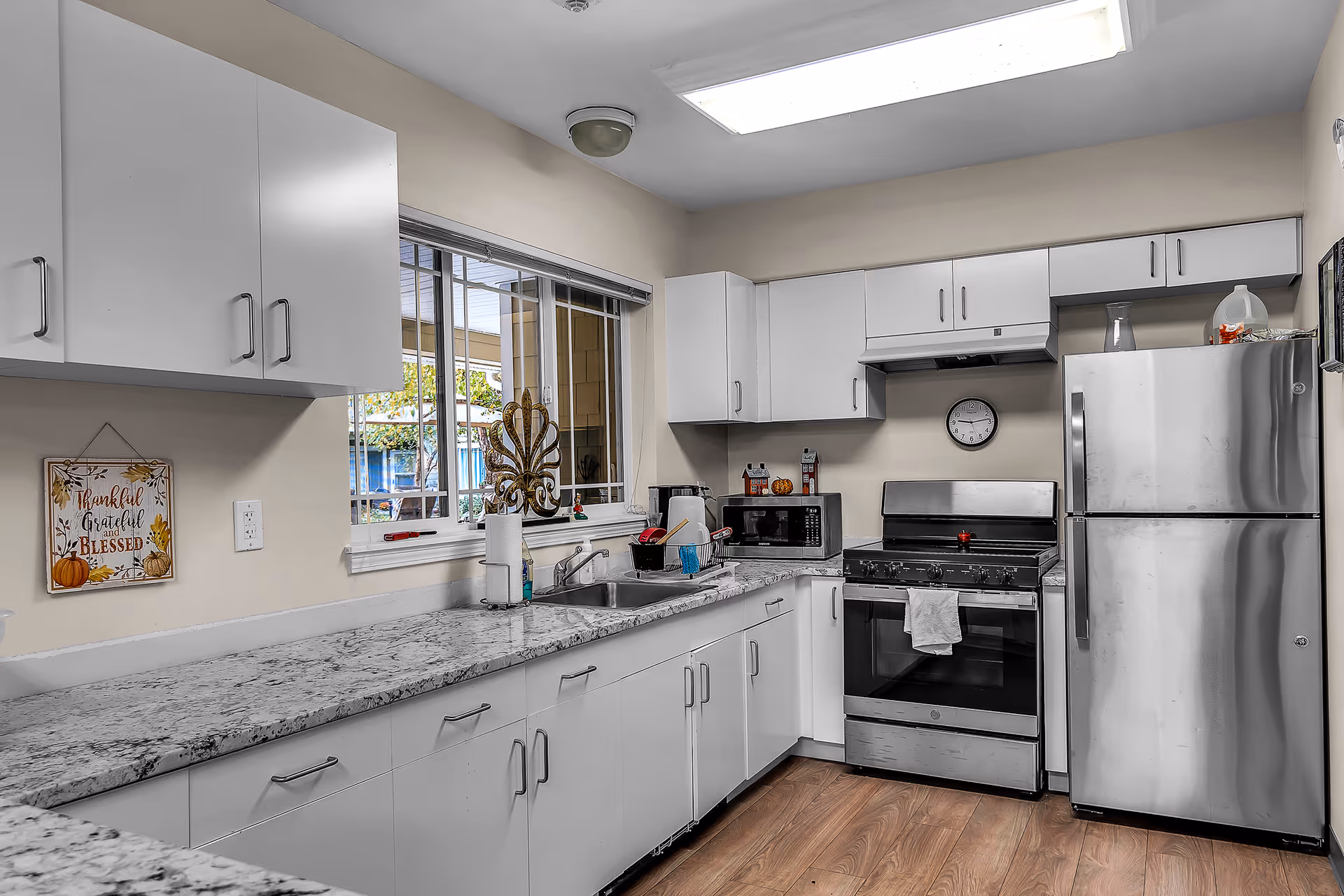 A modern kitchen with white cabinets, a granite countertop, a stainless steel refrigerator, and a stainless steel stove with an oven. There is a window above the sink with a view of the outdoors, a microwave on the counter, and a clock on the wall above the stove. The floor is wooden, and there is a decorative sign on the wall that reads 'Thankful Grateful Blessed.'