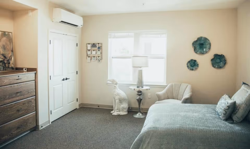 A softly lit bedroom with a bed, armchair, dresser, lamp by the window and decorative wall art including a white dog statue.