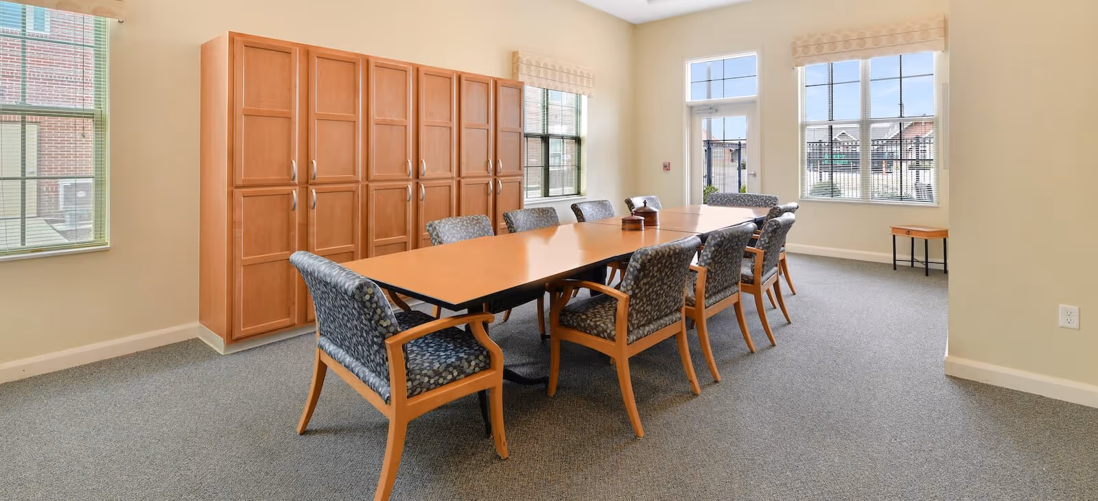 A bright room with a long rectangular wooden table surrounded by eight cushioned chairs with patterned upholstery. The room has beige walls, large windows letting in natural light, and a set of tall wooden cabinets along one wall. Outside the windows, a fenced outdoor area is visible.