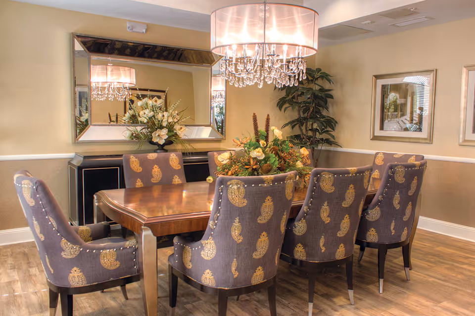 A dining room with a wooden table surrounded by six upholstered chairs with a paisley pattern. A large rectangular mirror hangs on the wall above a black sideboard with floral arrangements. A chandelier with crystal accents and a drum shade hangs above the table. The room has beige walls, framed artwork, and a potted plant in the corner.