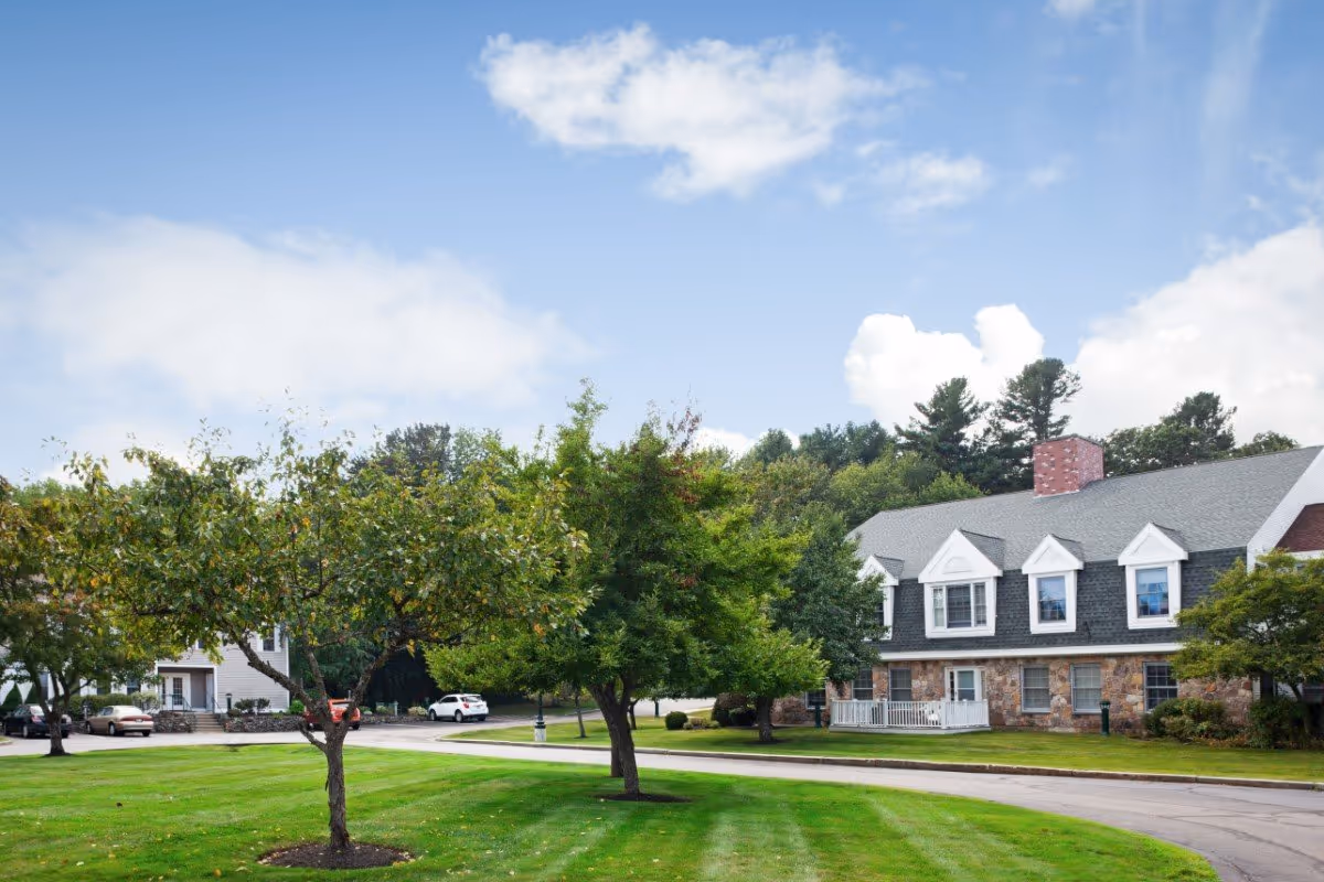 A well-maintained outdoor area of Huntington Common featuring a curved driveway, green lawn with neatly trimmed grass, several trees, and a large residential-style building with stone and siding exterior, multiple windows, and a chimney under a partly cloudy sky.