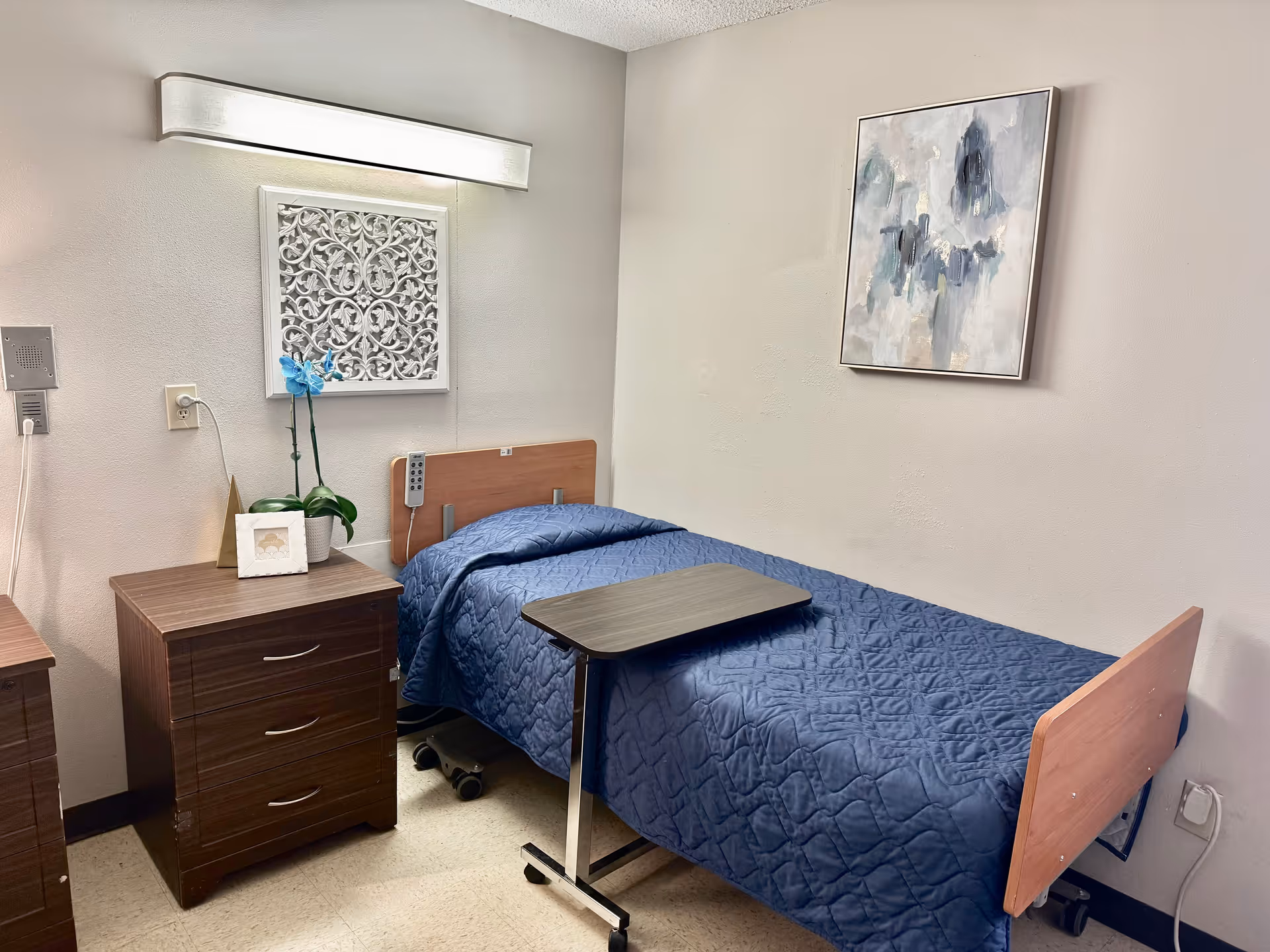 A small bedroom in a senior living facility with a single bed covered in a blue quilt. Next to the bed is a wooden nightstand with a potted plant and a framed photo. Above the bed is a decorative wall piece and a modern light fixture. A small overbed table is positioned on the bed. There is also an abstract painting hanging on the wall.