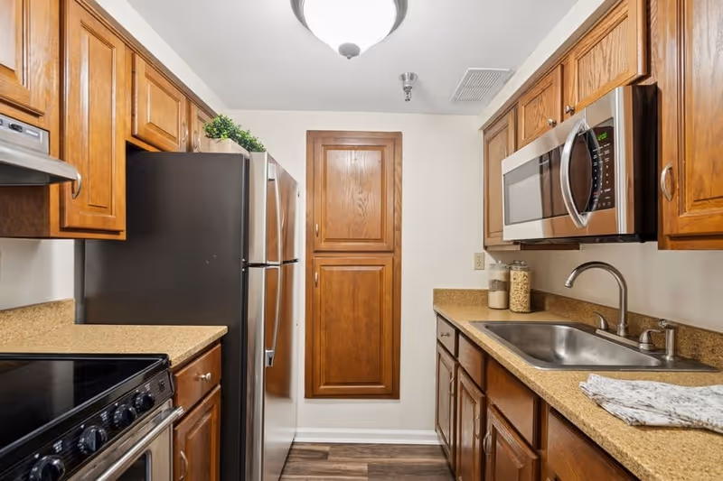 A compact kitchen with wooden cabinets, a stainless steel refrigerator, microwave, and stove. The countertop is beige with a sink on the right side and some jars and a folded cloth near the sink. The floor has a wood-like finish and the walls are painted white.