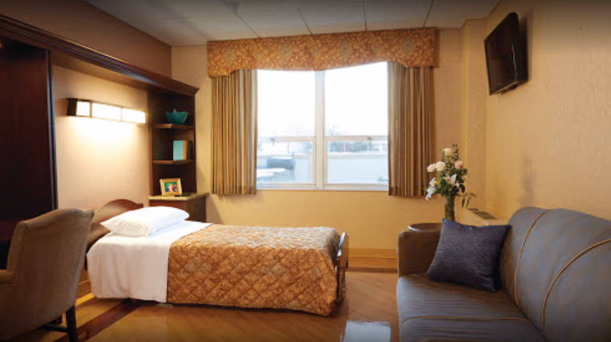 A cozy bedroom in a senior living facility featuring a single bed with a brown quilted bedspread, a cushioned chair, a small wooden shelf with decorative items, a window with beige curtains, a wall-mounted TV, a gray sofa with a dark pillow, and a vase of flowers on a side table.