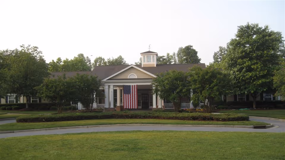 Front exterior view of a single-story building with a peaked roof and a small cupola on top. The entrance features white columns and a large American flag hanging in the center. The building is surrounded by green trees and well-maintained landscaping, with a circular driveway in front.