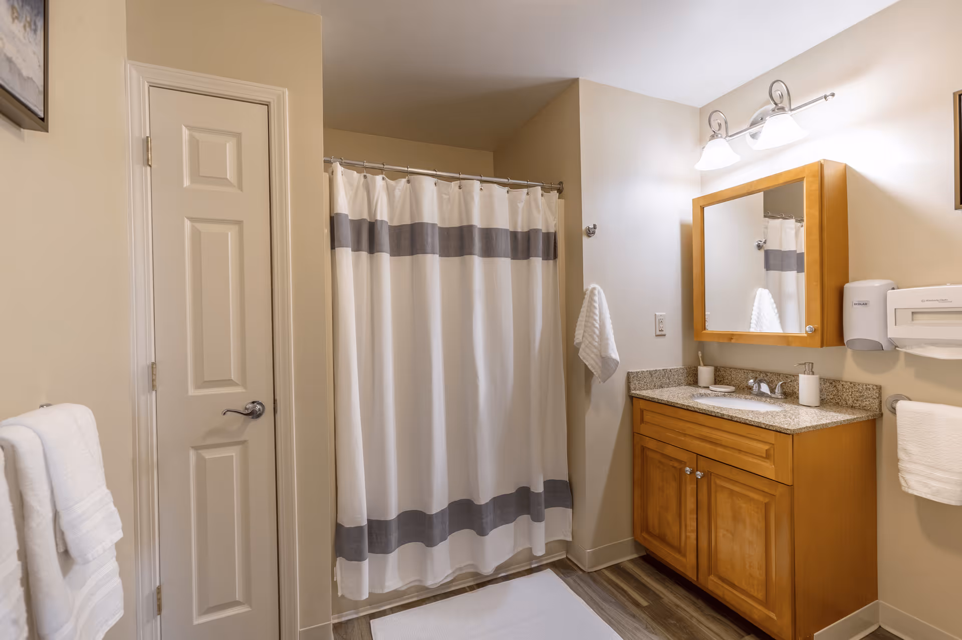 A clean bathroom with a shower curtain featuring gray stripes, a wooden vanity with a granite countertop, a mirror with wooden frame, wall-mounted light fixture, and white towels hanging on racks.