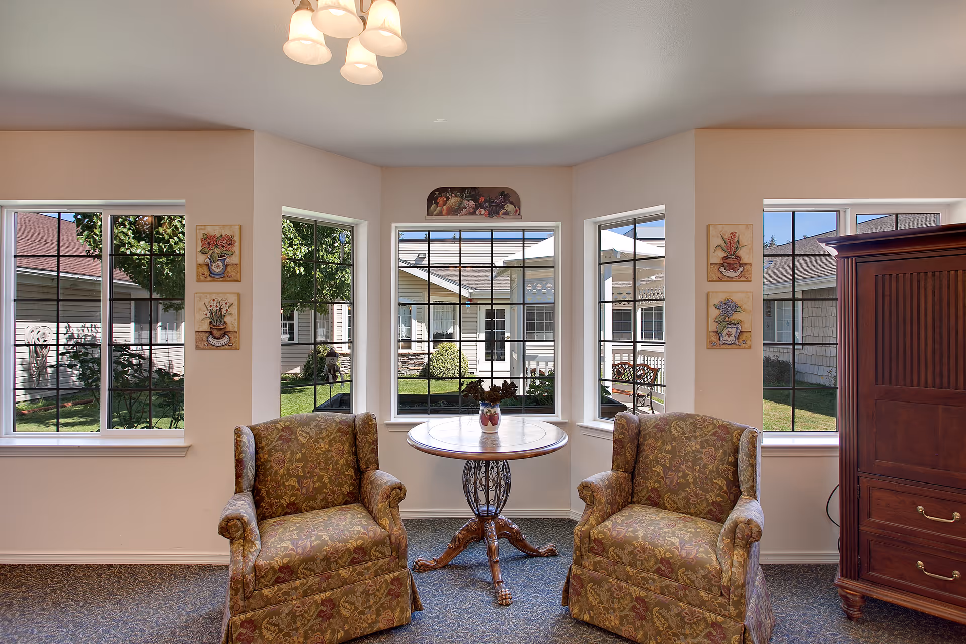 A cozy sitting area with two patterned armchairs facing a round wooden table with a small vase of flowers. The space is well-lit by natural light coming through five large windows that offer a view of a green courtyard with bushes and benches. The walls are decorated with small floral paintings, and there is a wooden cabinet on the right side.
