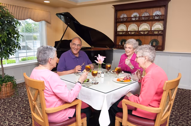 Four elderly residents sit around a dining table enjoying a meal in an elegant room with a grand piano and a china cabinet.