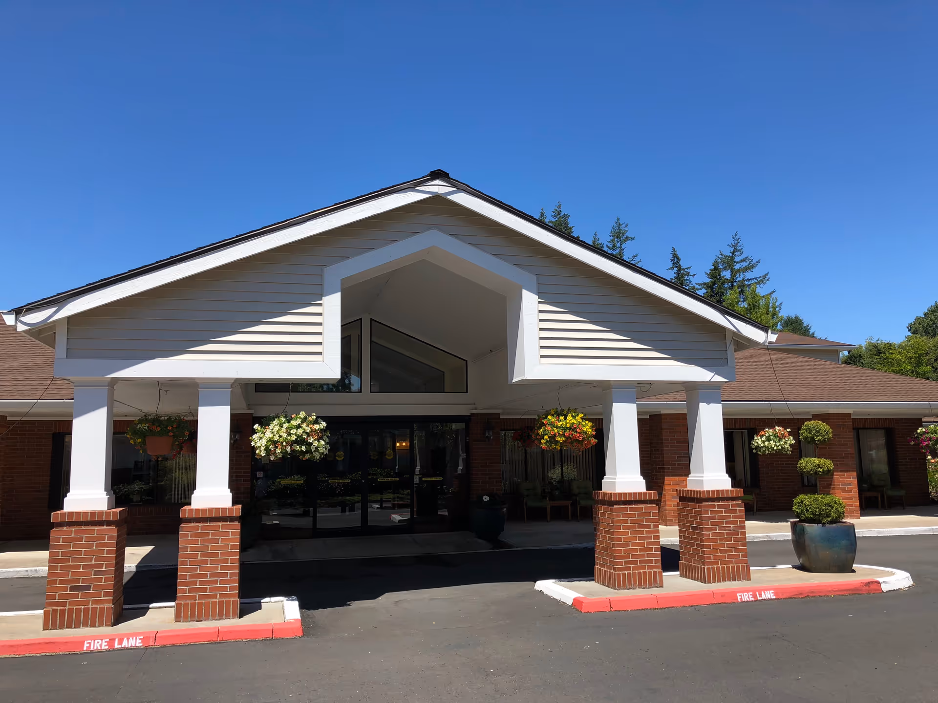 Front exterior view of Powell Valley Assisted Living and Memory Care building with a covered entrance supported by brick and white pillars, hanging flower baskets, and a clear blue sky.