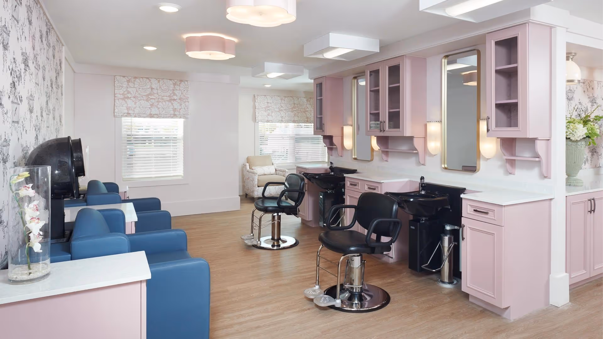 A bright and clean salon area in a senior living facility with pink cabinetry, black salon chairs, and wash basins. The room has wooden flooring, floral wallpaper on one wall, and windows with patterned blinds letting in natural light. There are blue armchairs and a white armchair in the background, along with decorative flowers in a glass vase.