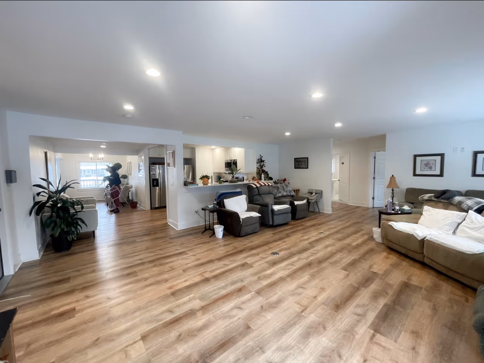 Spacious open living area with wood-look flooring, multiple sofas and recliners, plants, and an open kitchen in the background.