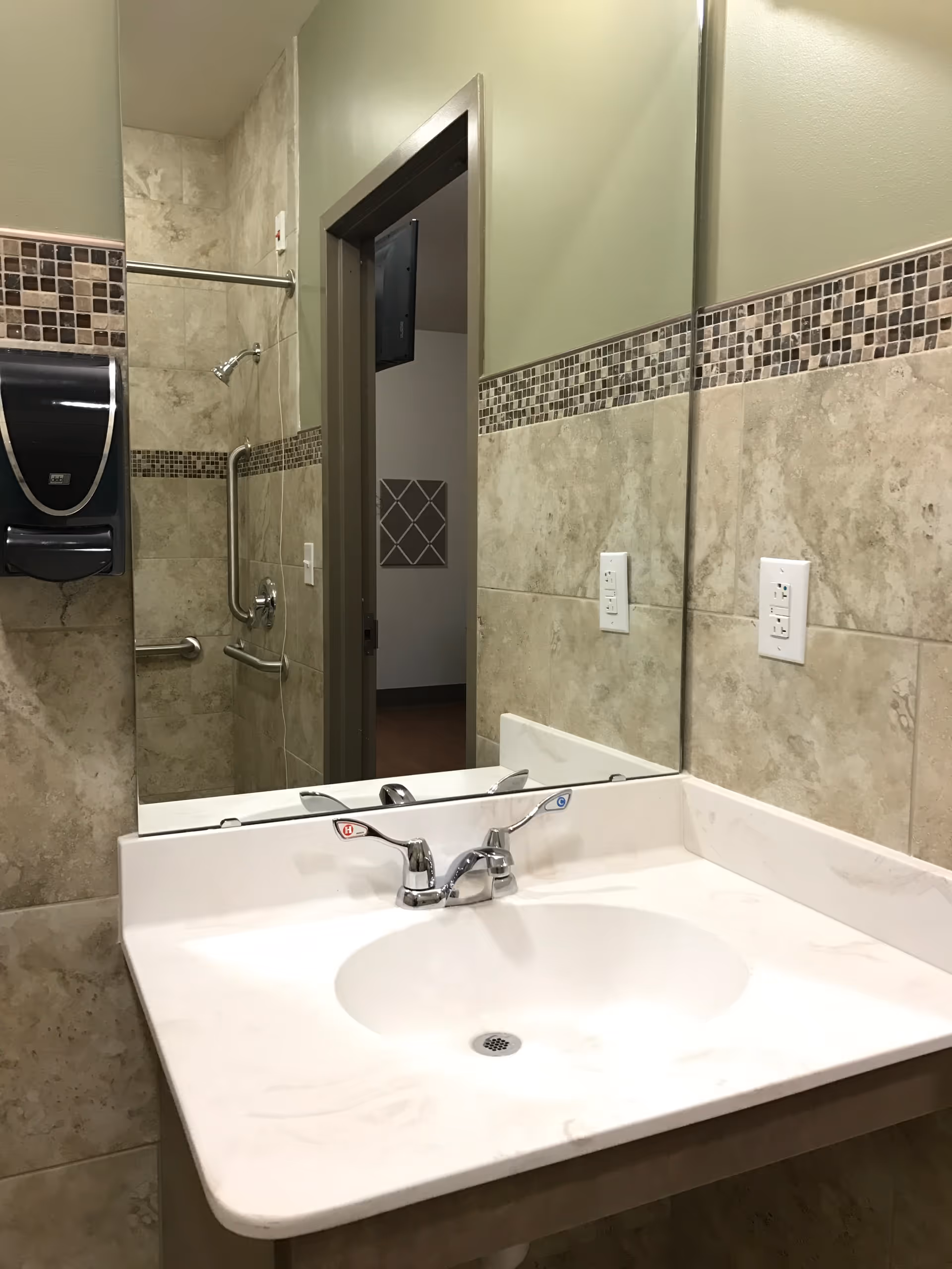 A bathroom sink with a white countertop and a chrome faucet with hot and cold handles. The wall behind the sink is tiled with beige tiles and a decorative mosaic tile border. A large mirror is mounted above the sink, reflecting a shower area with grab bars and a showerhead. A black paper towel dispenser is mounted on the left wall.