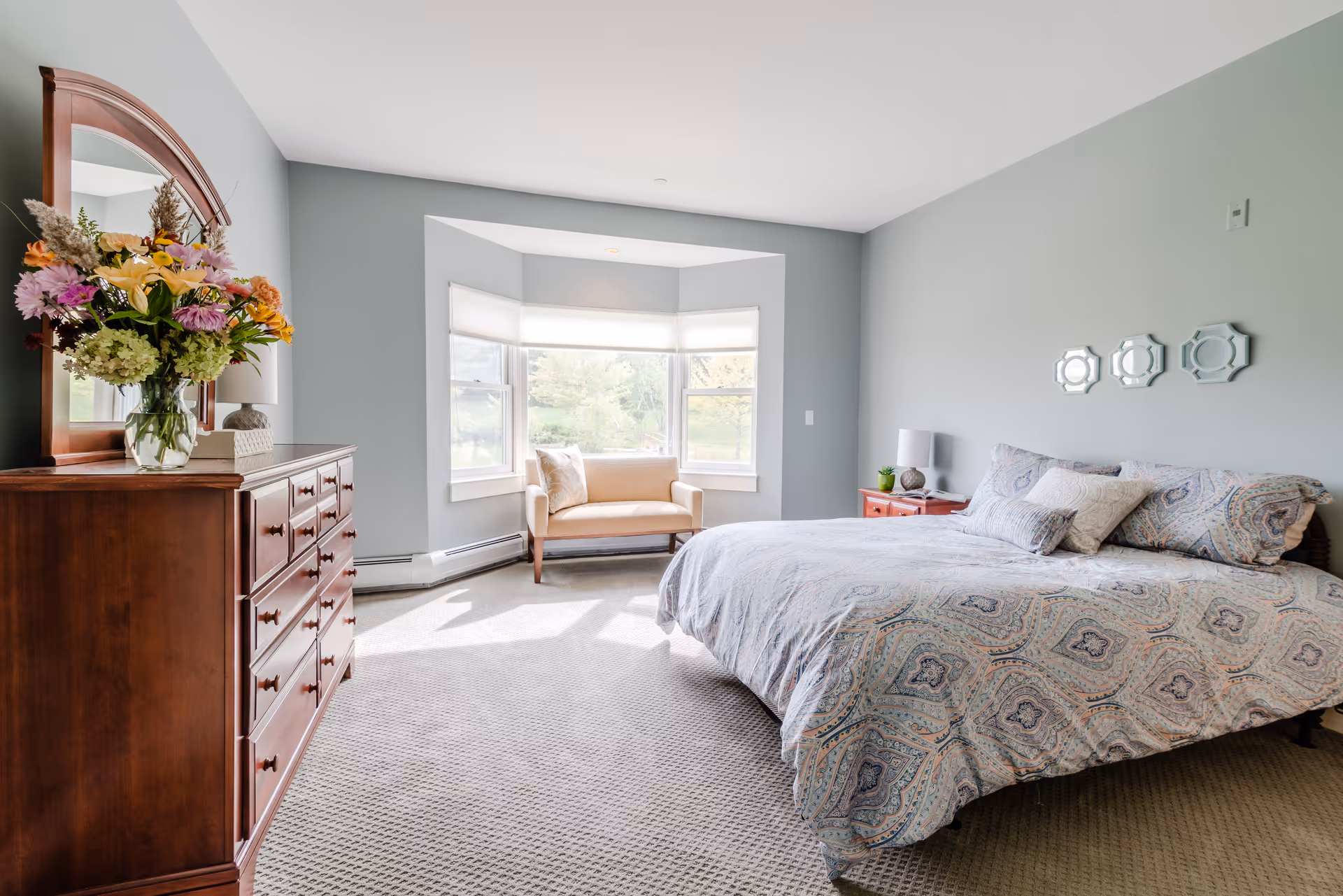 A bright and spacious bedroom with a large bed covered in patterned bedding, a wooden dresser with a mirror and a vase of colorful flowers, a small beige loveseat in front of a bay window with white blinds, and light gray walls.