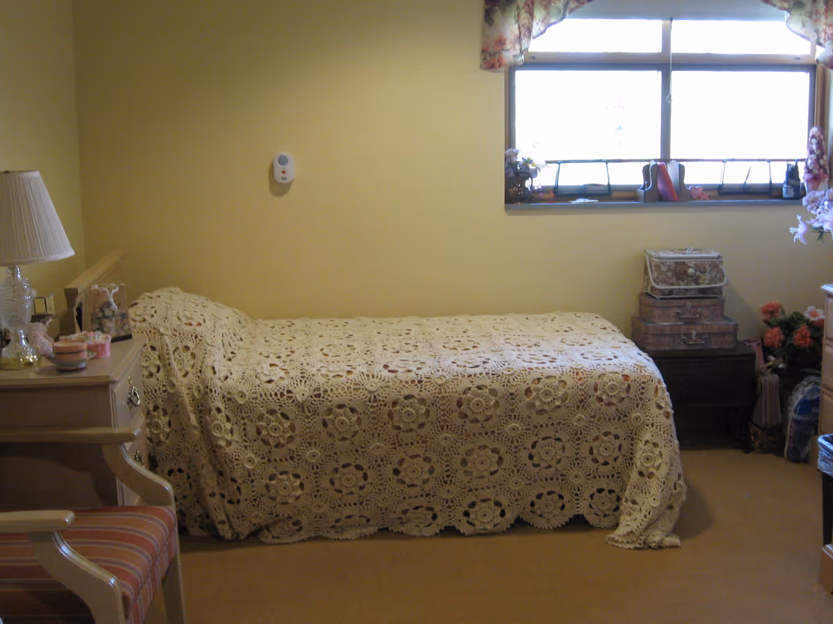 A small bedroom with a single bed covered by a cream-colored crocheted blanket. There is a window with floral curtains above the bed. Next to the bed is a nightstand with stacked decorative boxes and flowers. On the left side, there is a wooden chair with striped upholstery and a small table with a lamp and some decorative items.