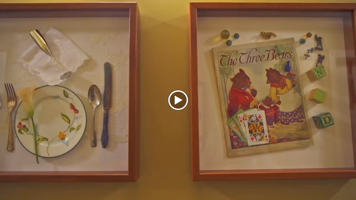 Two framed shadow boxes on a wall. The left frame contains a place setting with a plate, fork, knife, spoon, napkin, and a single flower. The right frame contains a vintage book titled 'The Three Bears' along with playing cards, toy blocks, marbles, and small figurines.