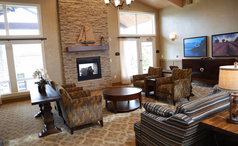 A cozy living room area with patterned armchairs and a striped sofa arranged around a round wooden coffee table. There is a stone fireplace with a wooden mantel holding a decorative sailboat model. Large windows and a glass door let in natural light. The walls have framed landscape artwork, and a wooden side table with a lamp is visible.
