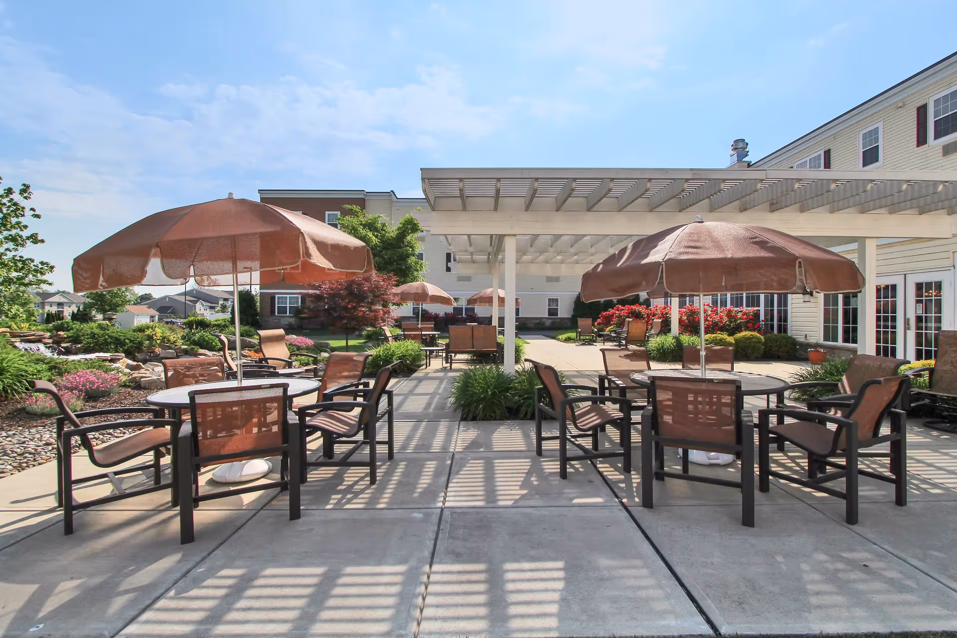 Outdoor patio area at Keystone Villa at Fleetwood with multiple round tables, each shaded by large brown umbrellas. The patio is surrounded by landscaped greenery, flowers, and a pergola providing partial shade. The building exterior is visible in the background under a clear blue sky.