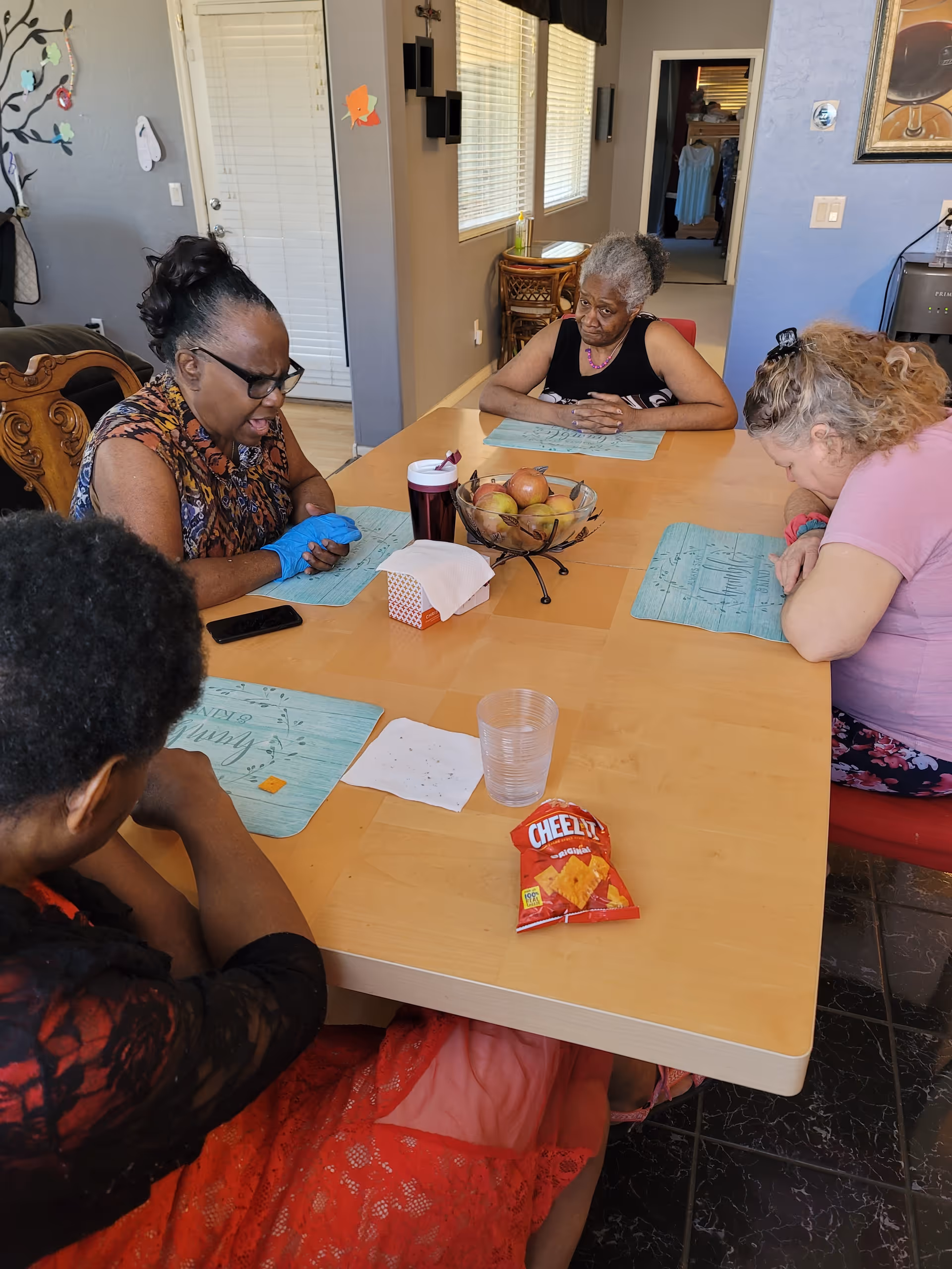 Four elderly women sitting around a wooden dining table in a well-lit room. The table has placemats, a bowl of fruit, a tissue box, a plastic cup, a packet of Cheez-It crackers, and a tumbler. The women appear to be engaged in a quiet activity or moment of reflection.