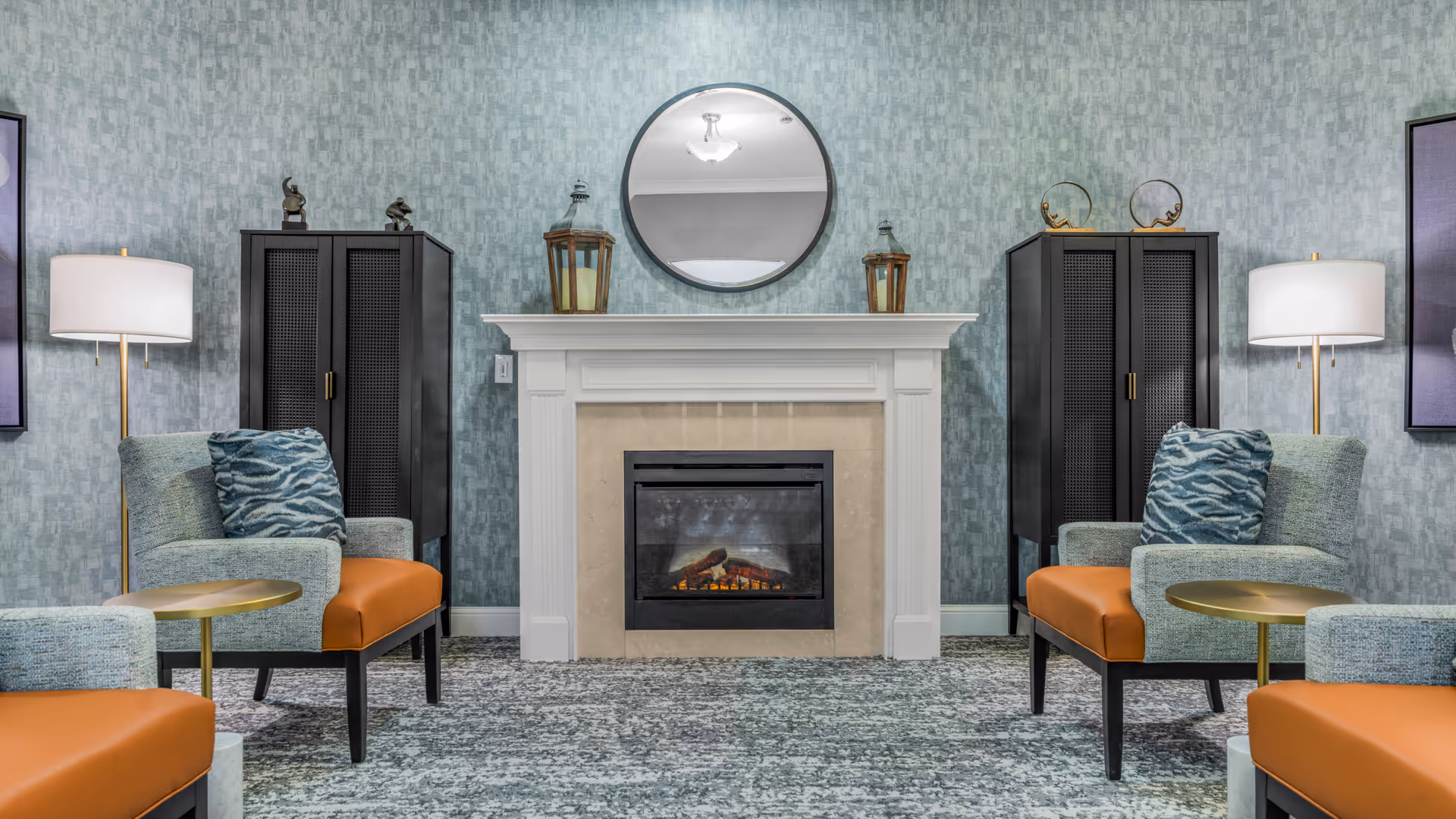 A cozy sitting area with a central electric fireplace surrounded by two black cabinets, each topped with decorative items. Two armchairs with orange seats and patterned blue cushions face each other, accompanied by small round gold side tables. The walls have a textured blue-gray wallpaper, and two floor lamps with white shades provide soft lighting. A round mirror hangs above the fireplace.