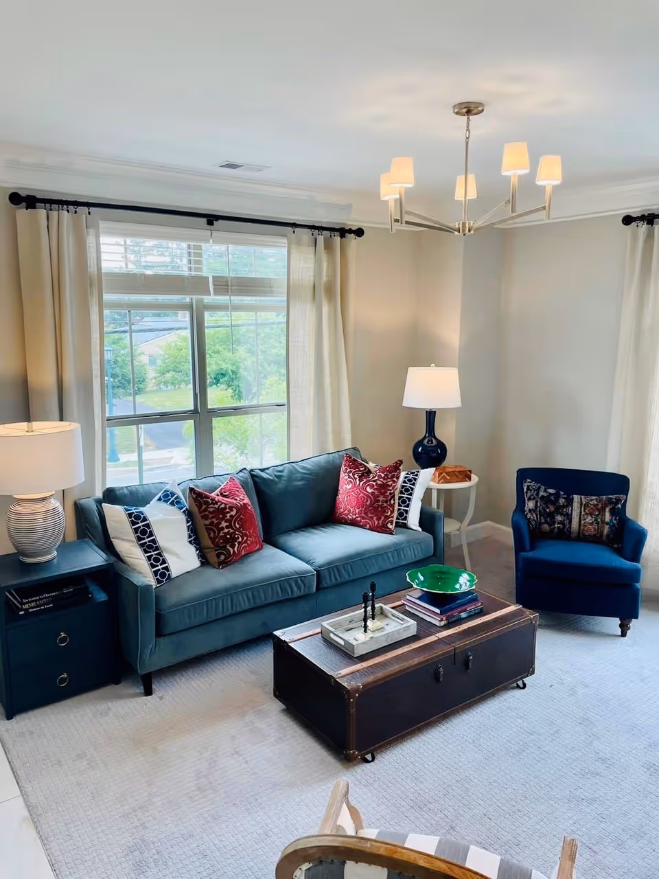 Bright living room with a teal sofa, blue armchair, trunk coffee table, lamps, and a large window with curtains.