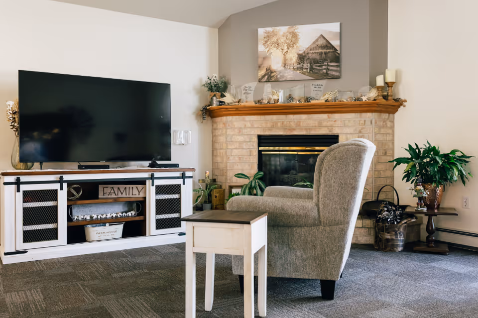 A cozy living room featuring a large flat-screen TV on a white wooden media console with decorative items including a sign that says 'FAMILY'. In front of the TV is a comfortable gray armchair and a small wooden side table. The room has a brick fireplace with a wooden mantel decorated with candles, plants, and framed artwork depicting a rustic barn scene. There are additional plants and decorative baskets around the fireplace.